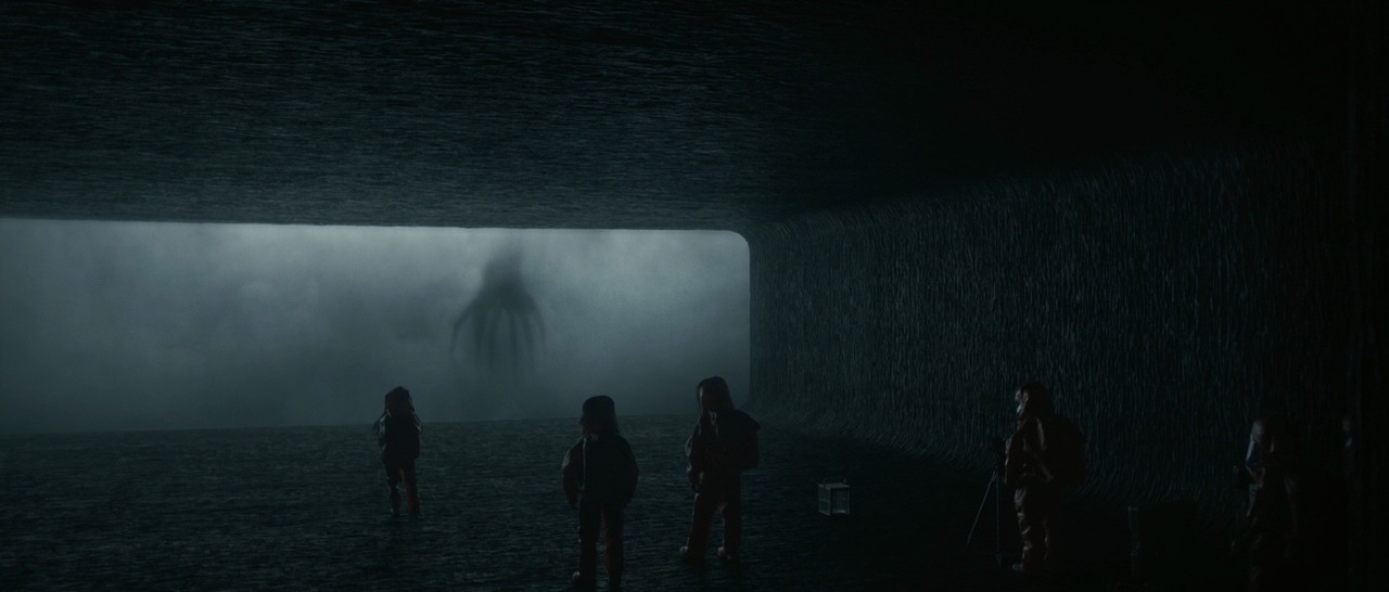 A dark viewing chamber with several people in silhouette facing a huge bright window, where a shadowy seven-limbed alien looms in the mist beyond.