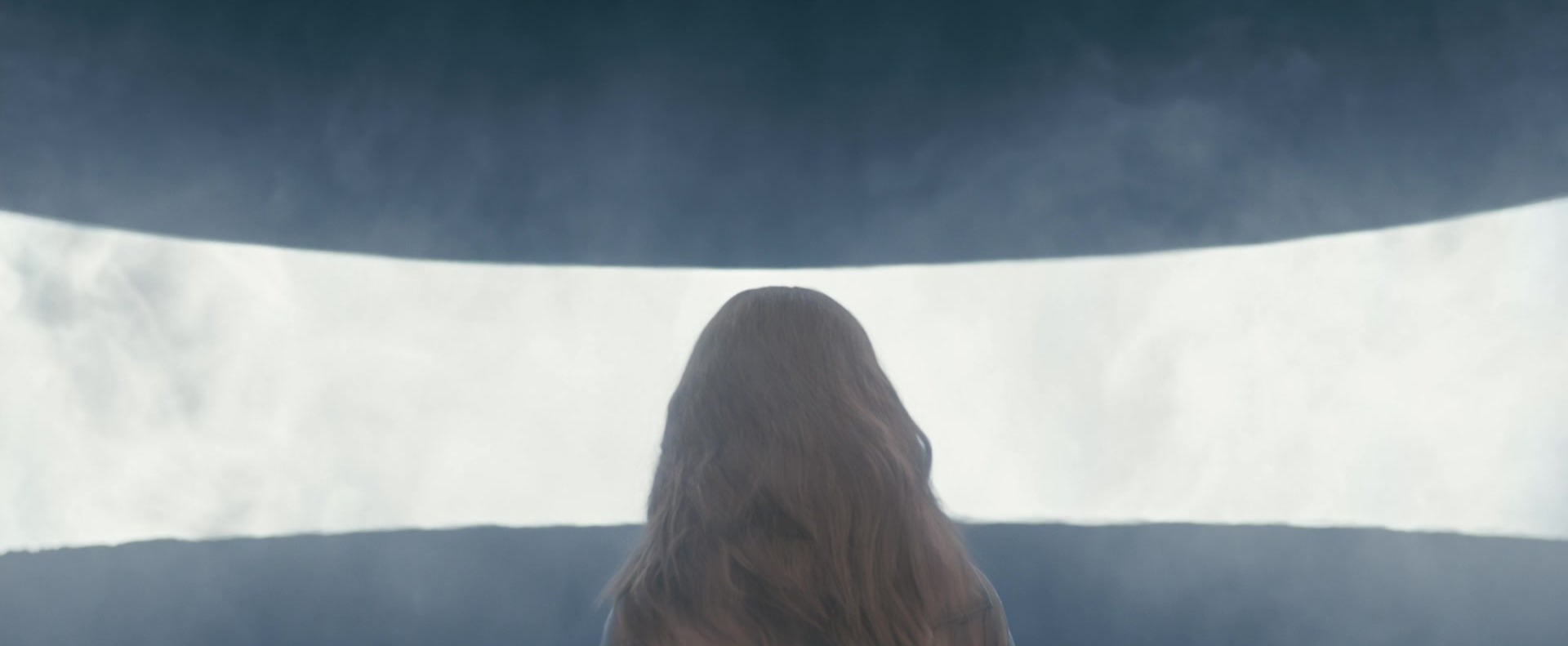 The back of a long-haired woman facing a wide horizontal band of bright white light cutting across a dark curved wall.